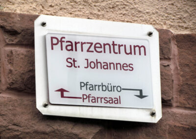 Wegweiser zu Pfarrbüro (jetzt unten) und Pfarrsaal (jetzt oben). Bei Veranstaltungen wird jedoch meist der barrierefreie Seiteneingang zum Pfarrsaal benutzt.